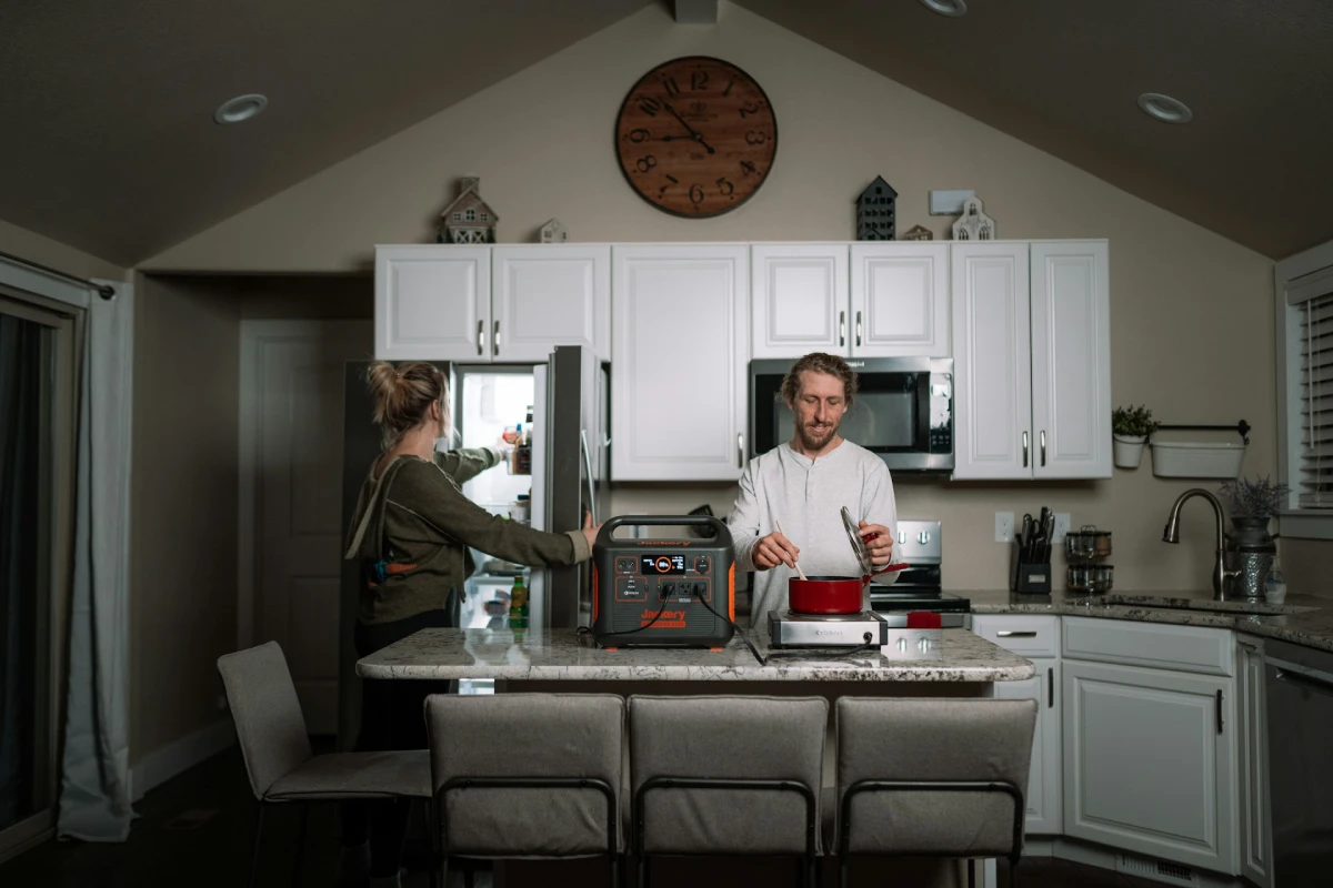 A kitchen home backup setup showing the Jackery Explorer 2000 v2 portable power station on a countertop while two people cook and organize food using fridge and stove appliances during a power outage scenario.